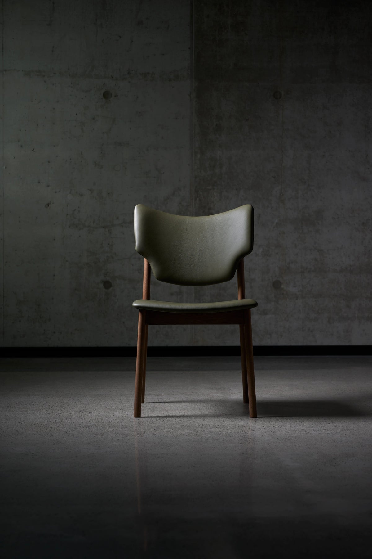 Front view of the dark green Eikund Veng Dining Chair in a dim interior, showing the wing-shaped back, open gap above the seat, and narrow wood frame.
