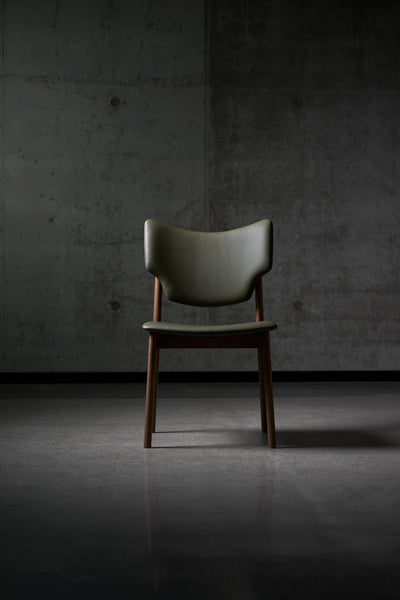 Front view of the dark green Eikund Veng Dining Chair in a dim interior, showing the wing-shaped back, open gap above the seat, and narrow wood frame.