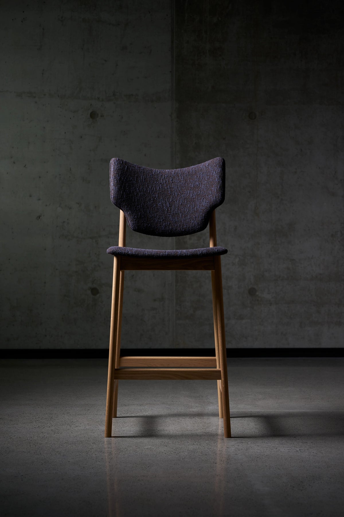 Front view of the dark purple Eikund Veng Bar Stool in a dim interior, showing its curved back with upturned corners, narrow seat, tall wood legs, and horizontal footrest.