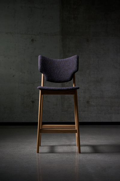 Front view of the dark purple Eikund Veng Bar Stool in a dim interior, showing its curved back with upturned corners, narrow seat, tall wood legs, and horizontal footrest.