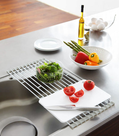 White Yamazaki Home Over-the-Sink Dish Drainer - Steel spanning a stainless-steel sink, supporting a cutting board with sliced tomatoes and a knife beside a glass bowl of greens.