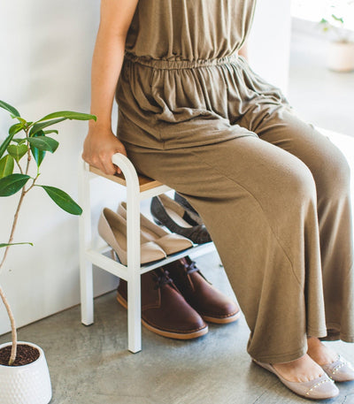 Person wearing a brown jumpsuit sitting on a chair with shoes underneath, in a room with a plant.