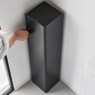 A person turns the empty black Yamazaki Home Rolling Bathroom Organizer - Steel in a bathroom corner, showing its tall rectangular panels and square top from above.