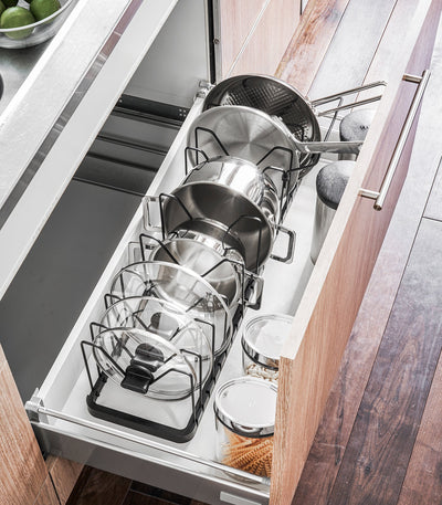Overhead view of the black Yamazaki Home Adjustable Pot Lid Organizer - Steel in a drawer, showing graduated cookware sizes and glass lids stored in separate upright slots.
