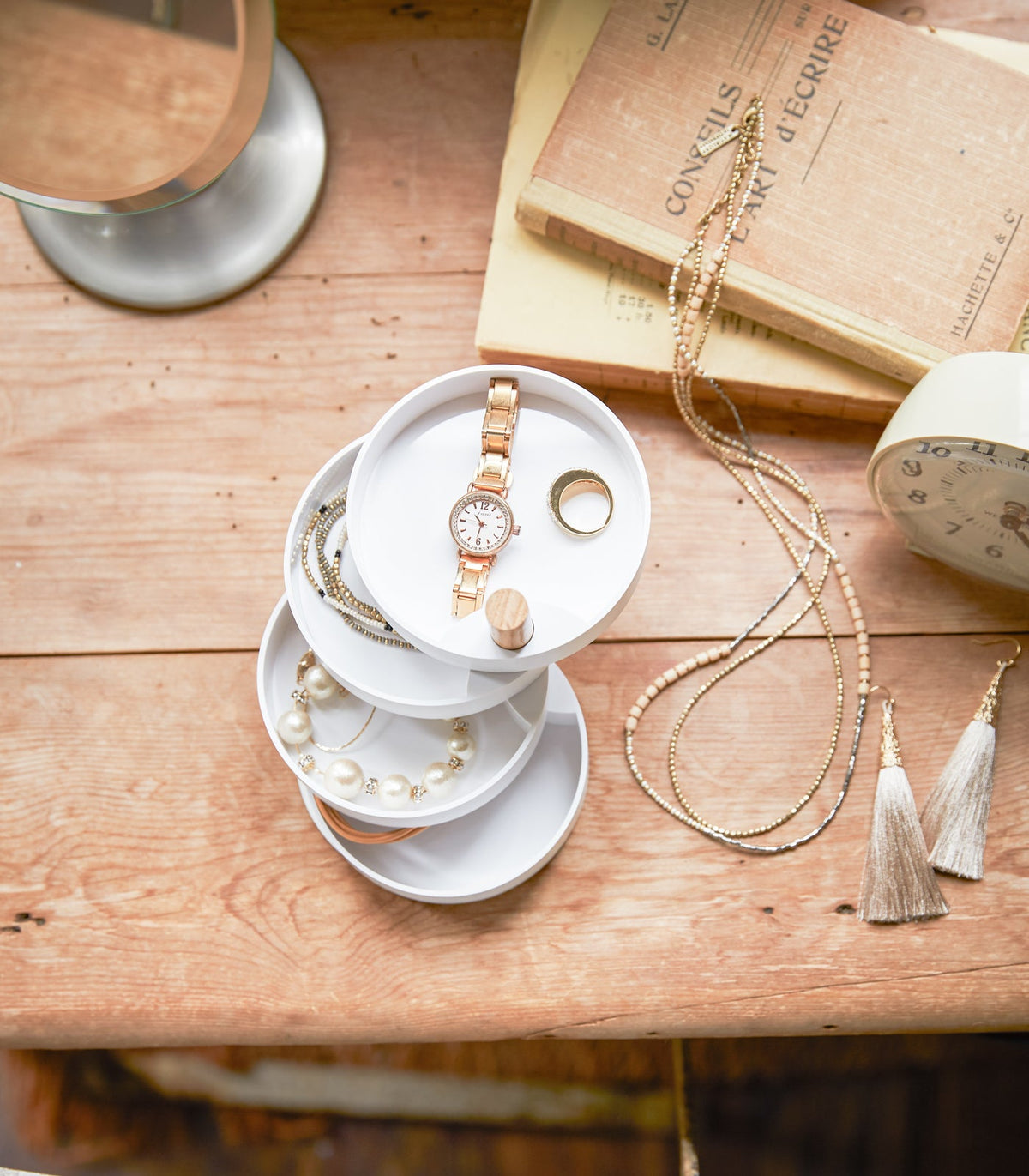 Overhead view of the Yamazaki Home Open Top Stacked Jewelry Organizer with trays partially spread across a wooden tabletop, holding a watch, ring and other jewellery beside books and long necklaces.