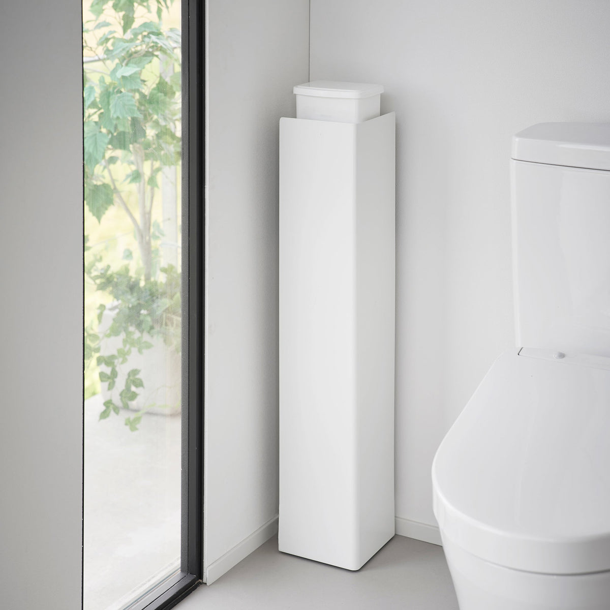 White Yamazaki Home Rolling Bathroom Organizer - Steel positioned with its solid side facing outward beside a toilet, concealing the storage compartments, with a white tissue box on top.