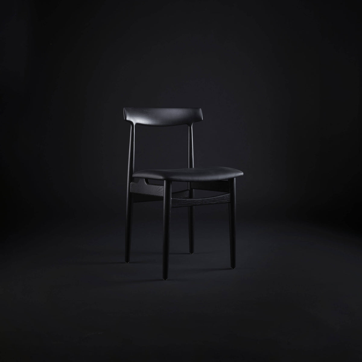 Black Eikund Hertug Dining Chair in a dark studio, shown from a three-quarter front angle with light highlighting the curved backrest and seat.