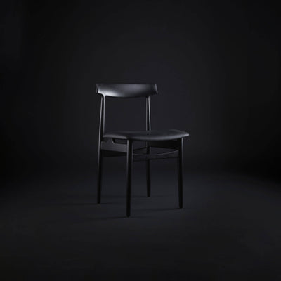 Black Eikund Hertug Dining Chair in a dark studio, shown from a three-quarter front angle with light highlighting the curved backrest and seat.