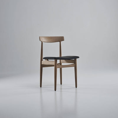 Three-quarter front view of the light wood Eikund Hertug Dining Chair with a black seat, showing the open back and curved rails beneath the seat.