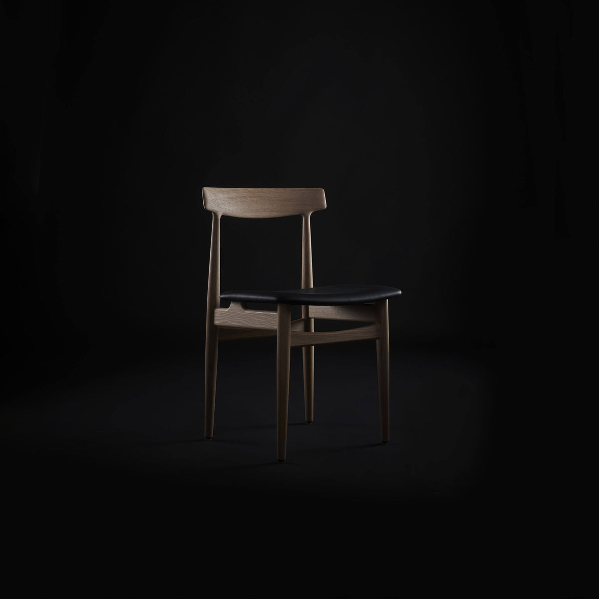 Light brown wood Eikund Hertug Dining Chair with a black seat in a dim studio, showing its narrow backrest and exposed frame under focused lighting.