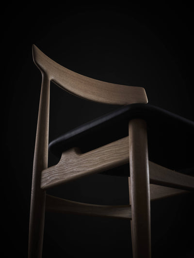 Low-angle close-up of the light wood Eikund Hertug Dining Chair, showing the curved grain-patterned backrest, black seat edge, and rounded front leg joined to the side rail.