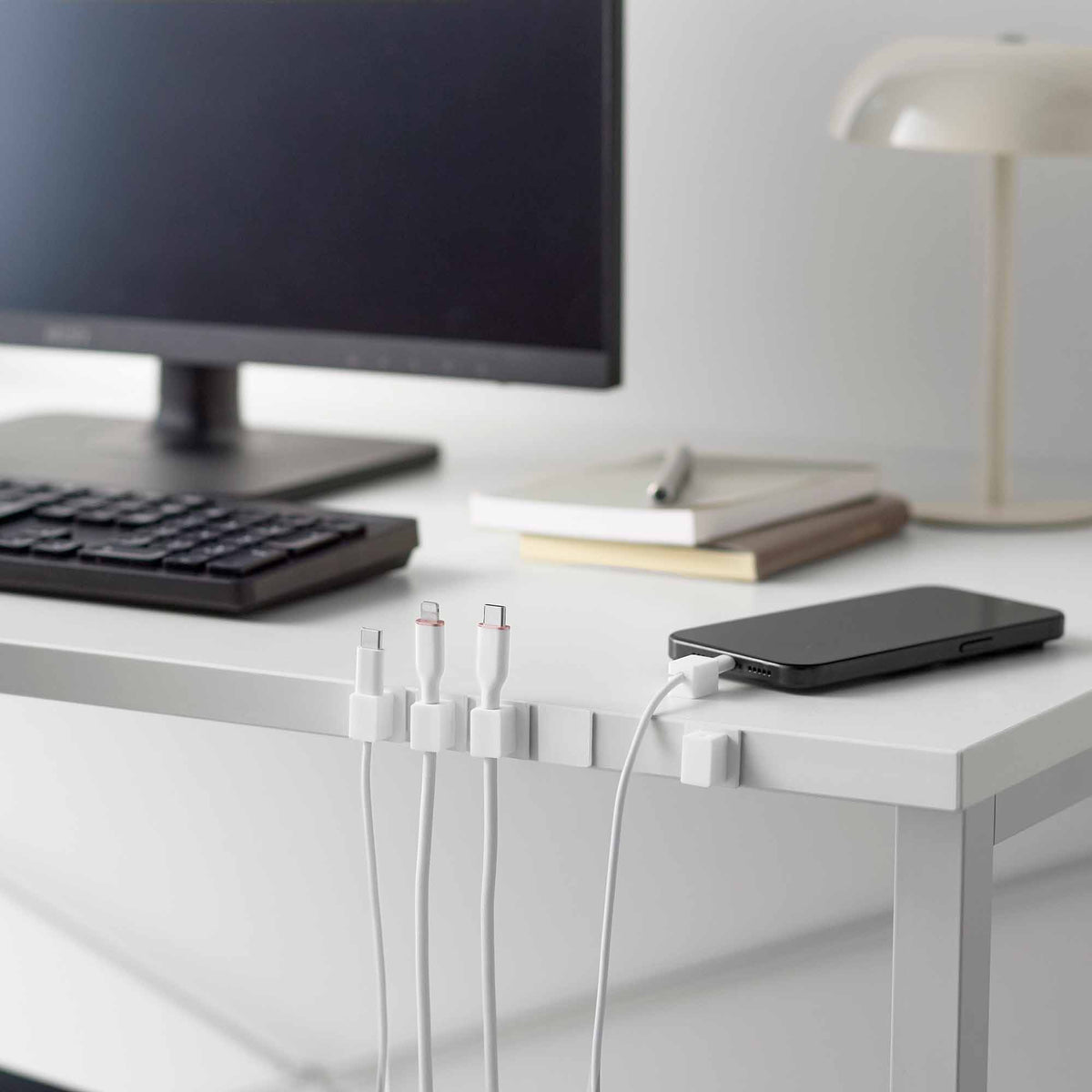 White Yamazaki Home Magnetic Cable Holders With Adhesive Steel Bases (Set of 5) - Silicone holding cables along a white desk beneath a monitor, beside a keyboard, lamp, books and phone.