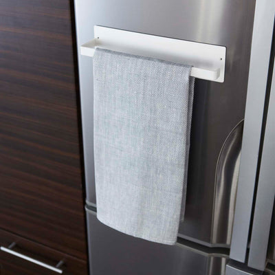 White Yamazaki Home Magnetic Paper Towel Holder - Steel holding a single folded grey kitchen towel against a silver refrigerator, viewed at an angle beside dark wood cabinetry.
