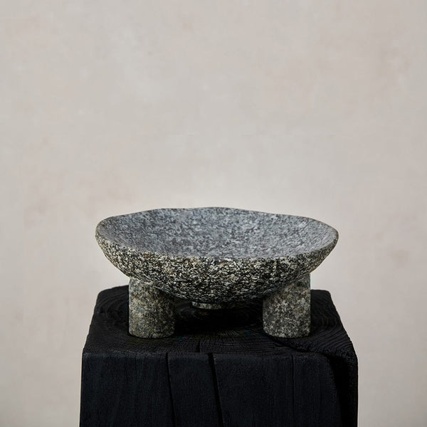 A carved stone footed bowl displayed on a black stand against a beige background.