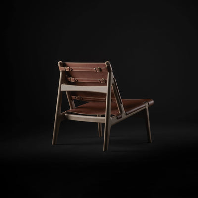 Three-quarter rear view of the tan leather Eikund Hunter Lounge Chair against black, showing buckled back straps, exposed wood crossbars, and the sloping seat frame.