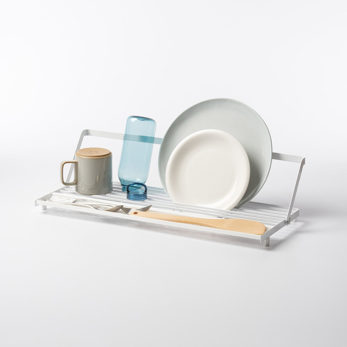 White Yamazaki Home Slim Over-the-Sink Dish Drying Rack - Steel holding two upright plates, a blue bottle, a lidded mug, cutlery and a wooden spatula, showing its narrow slatted base and raised support rail.