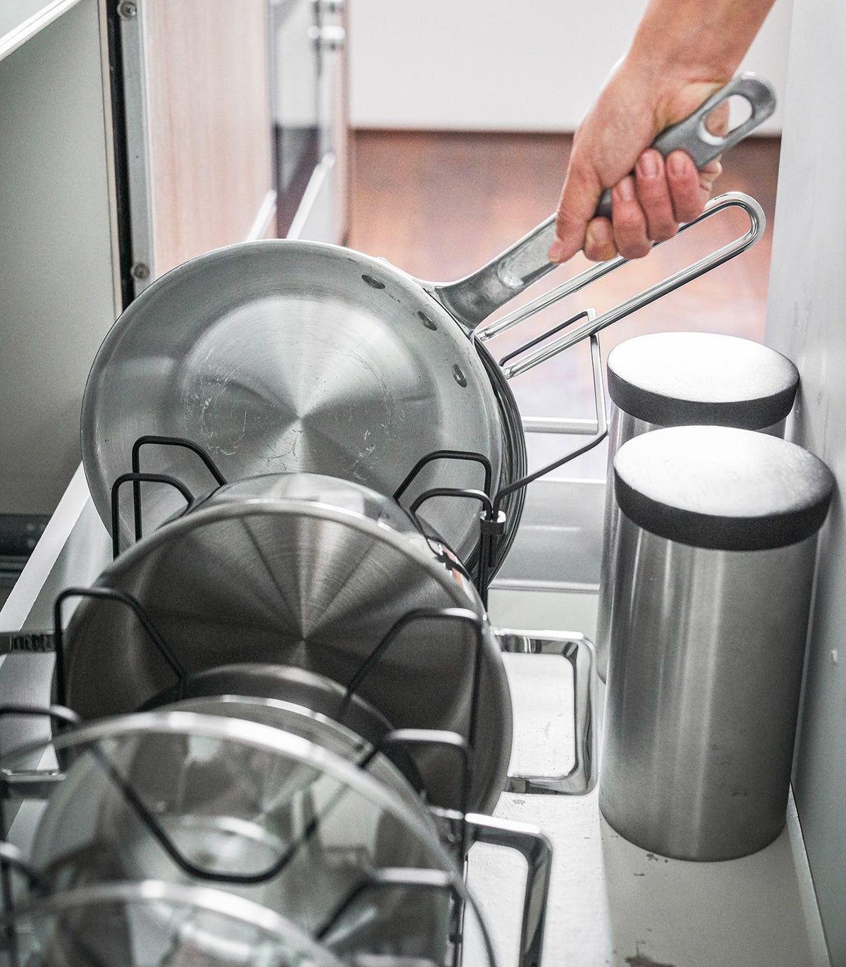 Close-up of silver-toned frying pans supported by the black Yamazaki Home Adjustable Pot Lid Organizer - Steel, showing the dividers beneath their long handles.