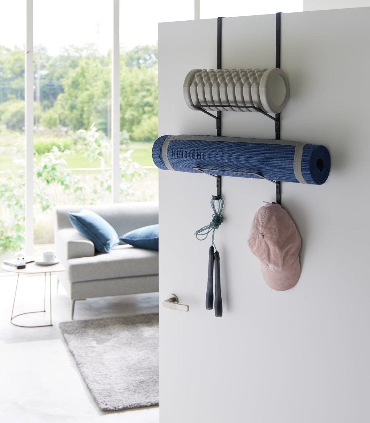 Black Yamazaki Home Over-the-Door Fitness Equipment Hanger - Steel on an open white door, holding a grey foam roller, rolled blue exercise mat, turquoise jump rope and pink cap in a bright living room.