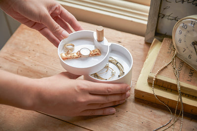 Two hands swivel the white trays of the Yamazaki Home Open Top Stacked Jewelry Organizer on a wooden dressing table, revealing a watch in the upper tray and bracelets in the tray beneath.