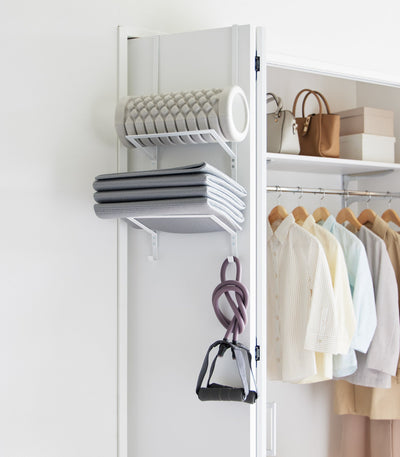 White Yamazaki Home Over-the-Door Fitness Equipment Hanger - Steel on a closet door, storing a grey foam roller, folded grey exercise mat and purple resistance bands with black handles.