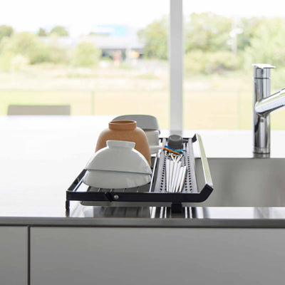 End view of the black Yamazaki Home Slim Over-the-Sink Dish Drying Rack - Steel over a sink, holding pale bowls and brown cups beside the narrow utensil tray.