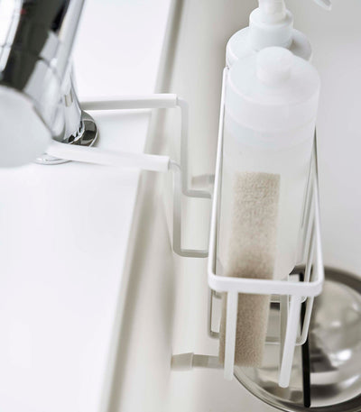 Side close-up of the white Yamazaki Home Faucet-Hanging Sponge Caddy - Steel, showing a sponge and tall white bottle inside the narrow basket and bent arms resting over the sink edge.