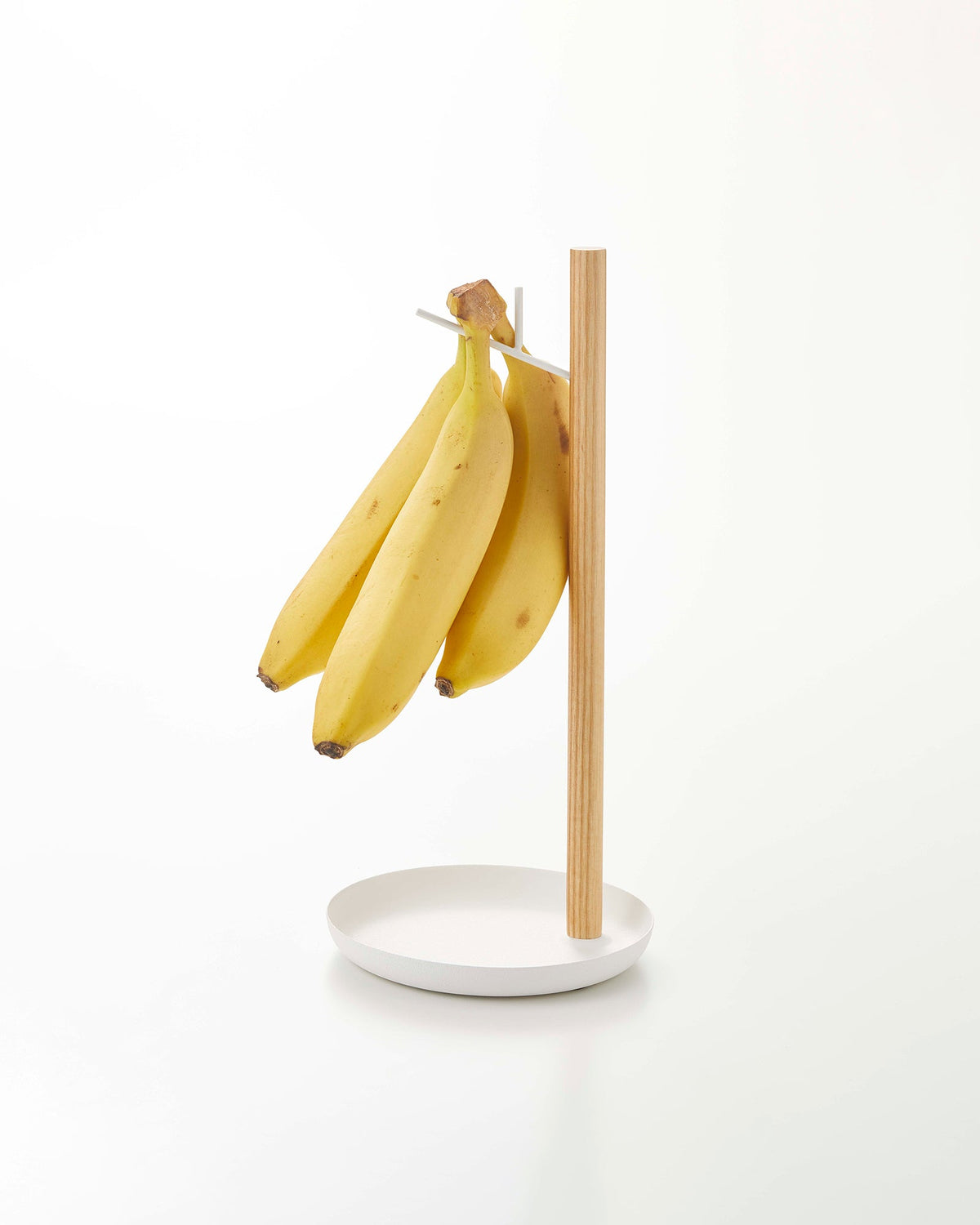 Yamazaki Home Banana Stand - Steel + Wood holding a bunch of yellow bananas on a white background, with a light wood upright, angled white hook and shallow circular white base.