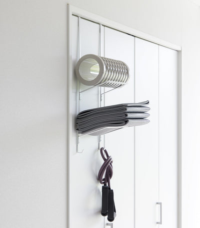 White Yamazaki Home Over-the-Door Fitness Equipment Hanger - Steel mounted on a white sliding closet door, holding a foam roller, folded grey mat and purple resistance bands.