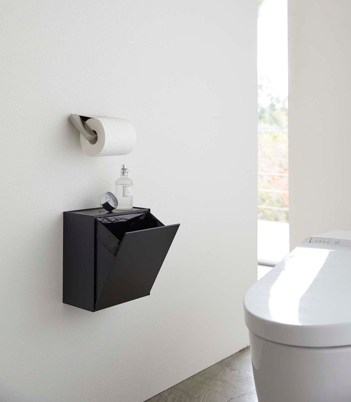 Black wall-mounted trash can in a bathroom setting with toilet and window.