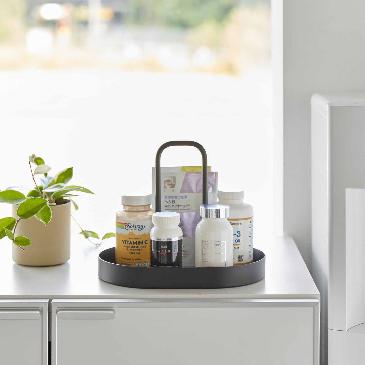 Front view of the black Yamazaki Home Vanity Tray with Handle - Steel holding bottles and upright packets beside a small green plant on a white cabinet.