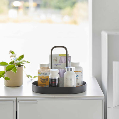 Front view of the black Yamazaki Home Vanity Tray with Handle - Steel holding bottles and upright packets beside a small green plant on a white cabinet.