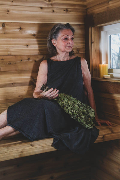 Merino wool sauna dress paired with fresh sauna greenery in a traditional wood sauna setting.