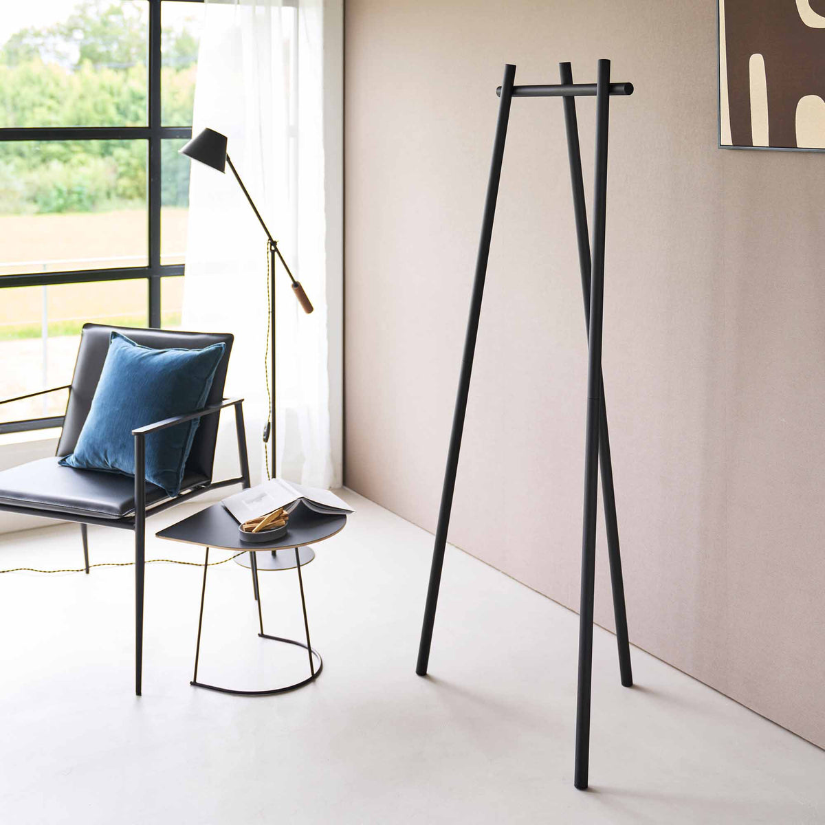 Empty black Yamazaki Home Tri-Post Coat Rack (60" H) - Steel in a sunlit room, showing three slim posts and a short top rail beside a chair, side table and floor lamp.