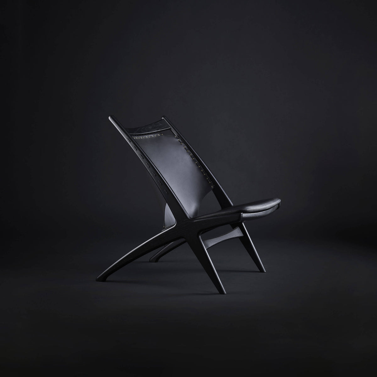 Side view of an all-black Eikund Krysset Lounge Chair in a dark studio, showing its sharply reclined back, thin seat, and distinctive X-shaped wood frame.