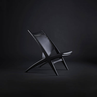 Side view of an all-black Eikund Krysset Lounge Chair in a dark studio, showing its sharply reclined back, thin seat, and distinctive X-shaped wood frame.