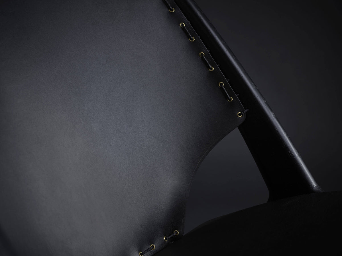 Close-up of the black Eikund Krysset Lounge Chair’s leather back panel, showing edge lacing beside the black wood upright and a curved opening near the seat.