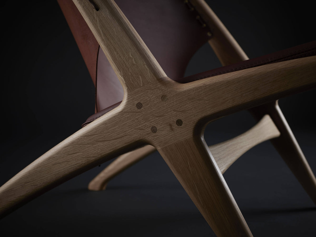 Close-up of the Eikund Krysset Lounge Chair’s crossed wood side frame, showing visible grain, round joinery details, and the angular junction beneath the leather back.