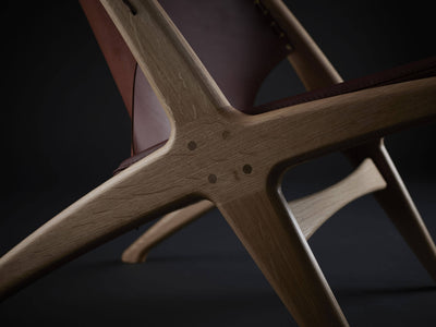 Close-up of the Eikund Krysset Lounge Chair’s crossed wood side frame, showing visible grain, round joinery details, and the angular junction beneath the leather back.