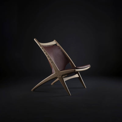 Eikund Krysset Lounge Chair with light wood and reddish-brown leather in a dark studio, shown at an angle to emphasize its laced back panel, slim seat, and crossed legs.
