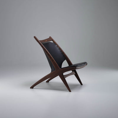 Three-quarter side view of the Eikund Krysset Lounge Chair with a warm brown wood frame and black leather, showing its reclined silhouette against a gray studio background.