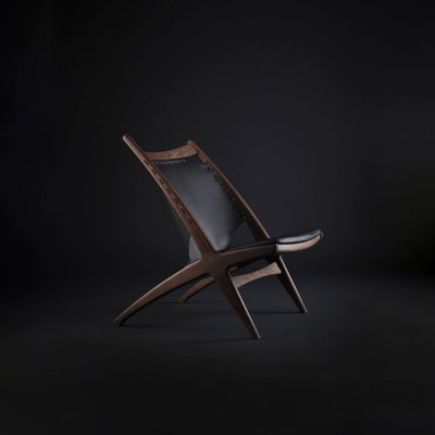 Warm brown wood and black leather Eikund Krysset Lounge Chair in a dark studio, showing the tall angled back, narrow cushioned seat, and crossed side frame.