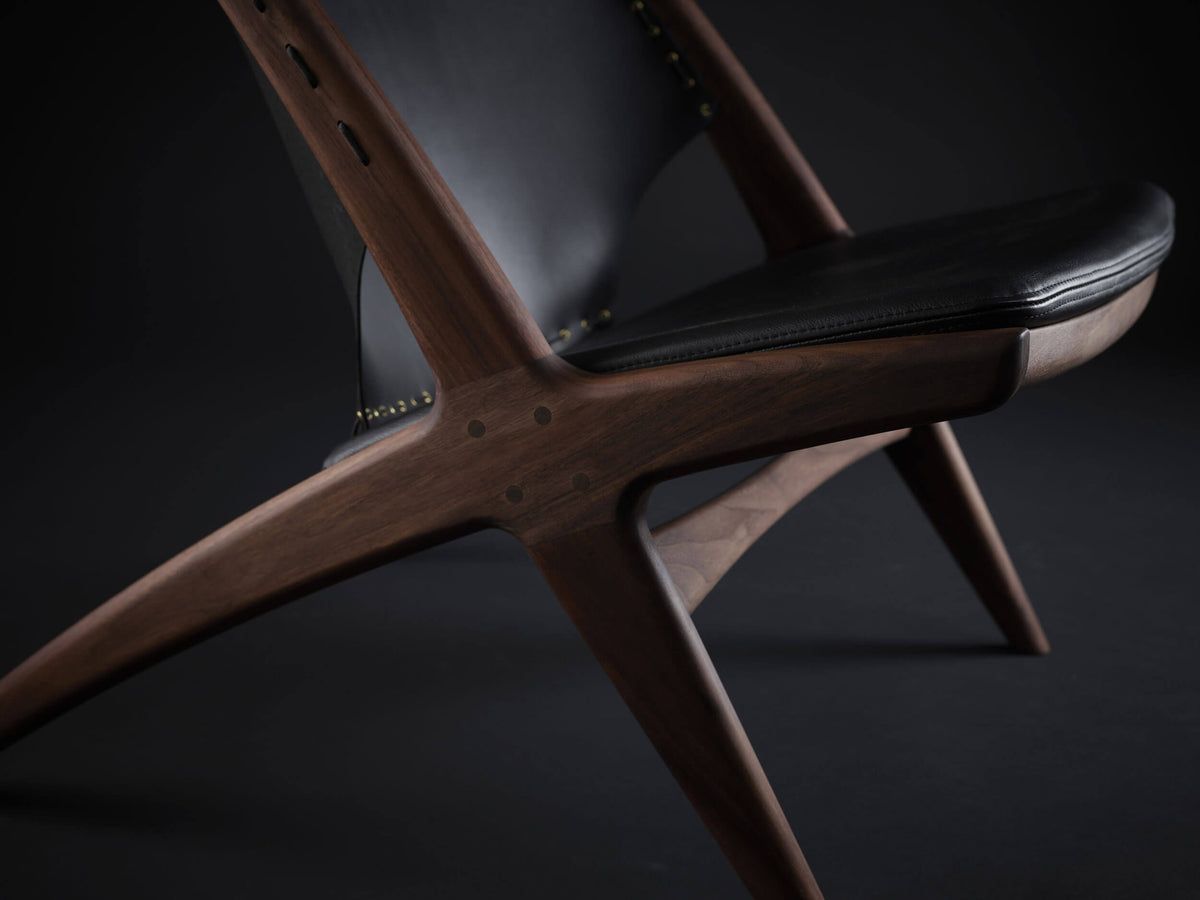Close-up of the Eikund Krysset Lounge Chair’s brown wood frame beneath the black leather seat and back, showing circular joinery details, visible grain, and intersecting supports.