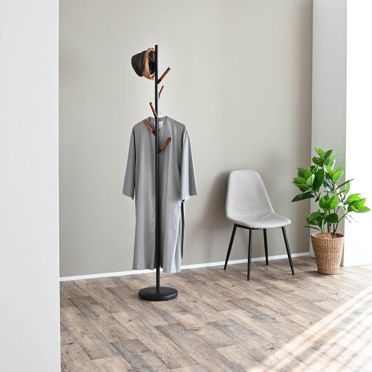 Black Yamazaki Home Coat Rack (70" H) - Steel holding a long grey coat and hat beside a grey chair and green potted plant on wood flooring.