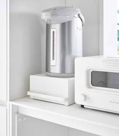 Silver electric kettle on a white shelf next to a white microwave.