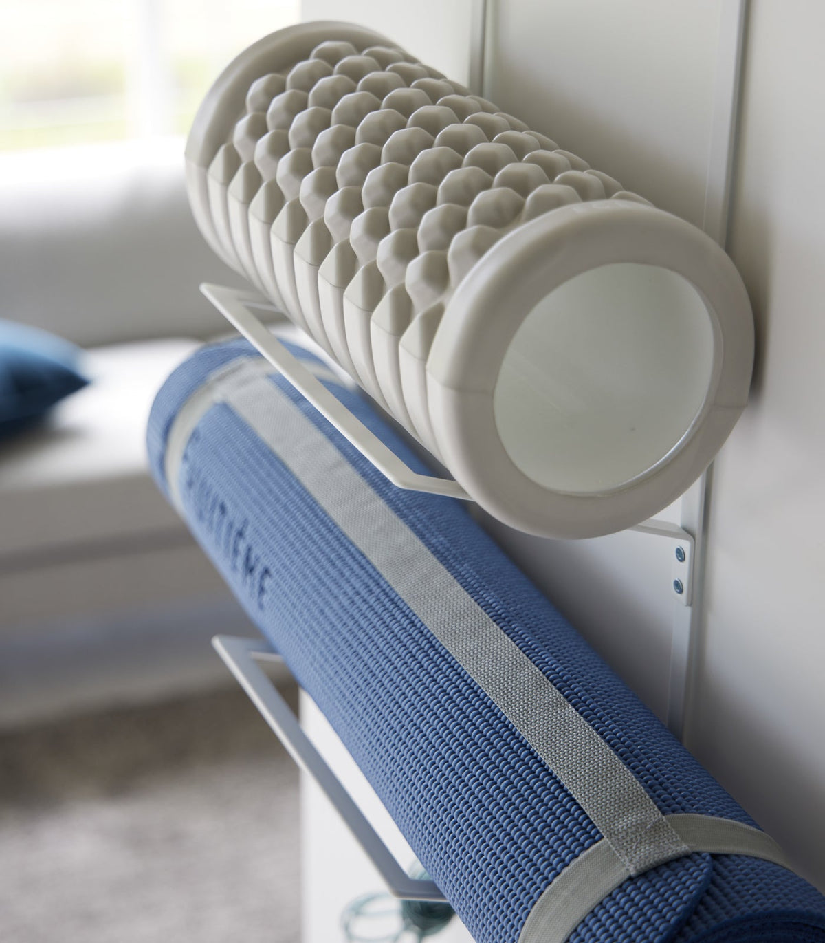 Side close-up of the white Yamazaki Home Over-the-Door Fitness Equipment Hanger - Steel, showing projecting supports cradling a textured grey foam roller and a strapped blue exercise mat.