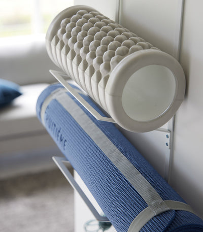 Side close-up of the white Yamazaki Home Over-the-Door Fitness Equipment Hanger - Steel, showing projecting supports cradling a textured grey foam roller and a strapped blue exercise mat.