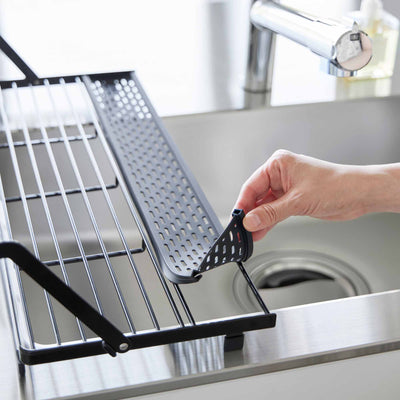 A hand lifts the black perforated utensil tray from the Yamazaki Home Slim Over-the-Sink Dish Drying Rack - Steel, showing the removable section above the sink.
