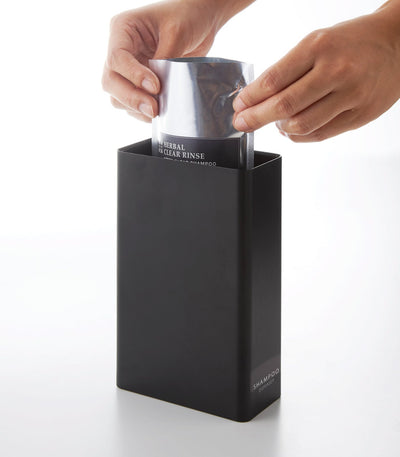 Two hands lower a refill pouch into the black Yamazaki Home Rectangle Shower Dispenser - Three Styles through its uncovered rectangular top.