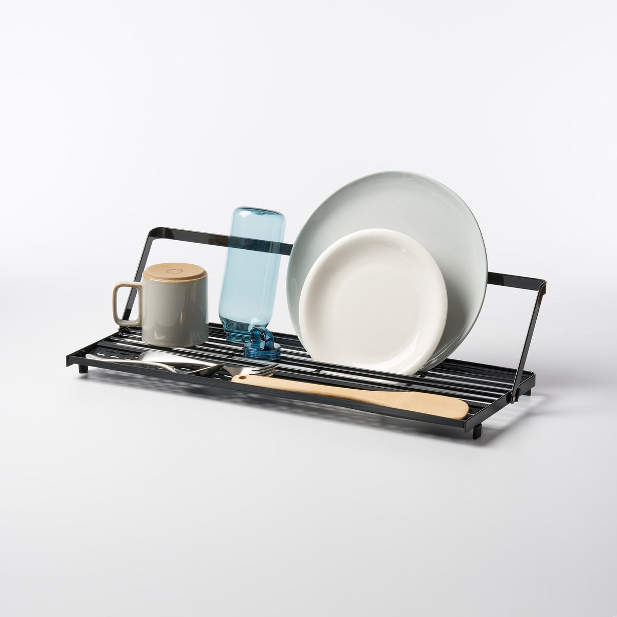 Black Yamazaki Home Slim Over-the-Sink Dish Drying Rack - Steel holding pale plates, a blue bottle, a lidded mug, cutlery and a wooden spatula against a light background.