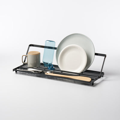 Black Yamazaki Home Slim Over-the-Sink Dish Drying Rack - Steel holding pale plates, a blue bottle, a lidded mug, cutlery and a wooden spatula against a light background.